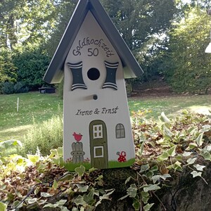 May include: A white wooden birdhouse with a gray roof and decorative accents. The birdhouse features the text "Goldhochzeit 50" and the names "Irene & Ernst". It is decorated with a painted bird, door, window, and mushroom.