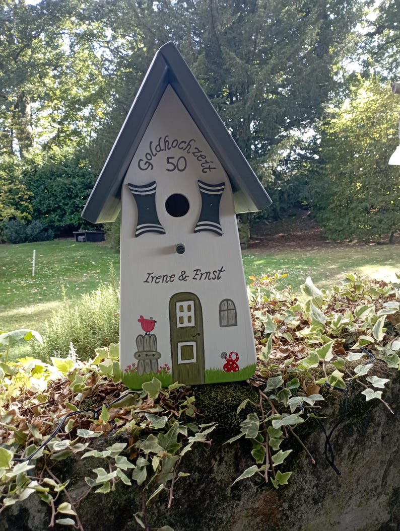 May include: A white wooden birdhouse with a gray roof and decorative accents. The birdhouse features the text "Goldhochzeit 50" and the names "Irene & Ernst". It is decorated with a painted bird, door, window, and mushroom.