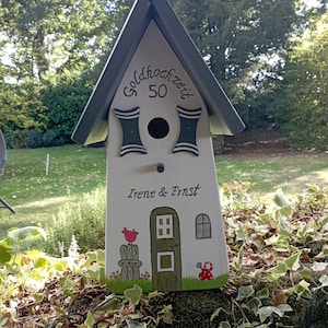 May include: A decorative birdhouse with a gray roof and white body. The birdhouse has the text "Goldhochzeit 50" and the names "Irene & Ernst". It features painted details like a door, window, and a red bird. The birdhouse is set against a backdrop of greenery.