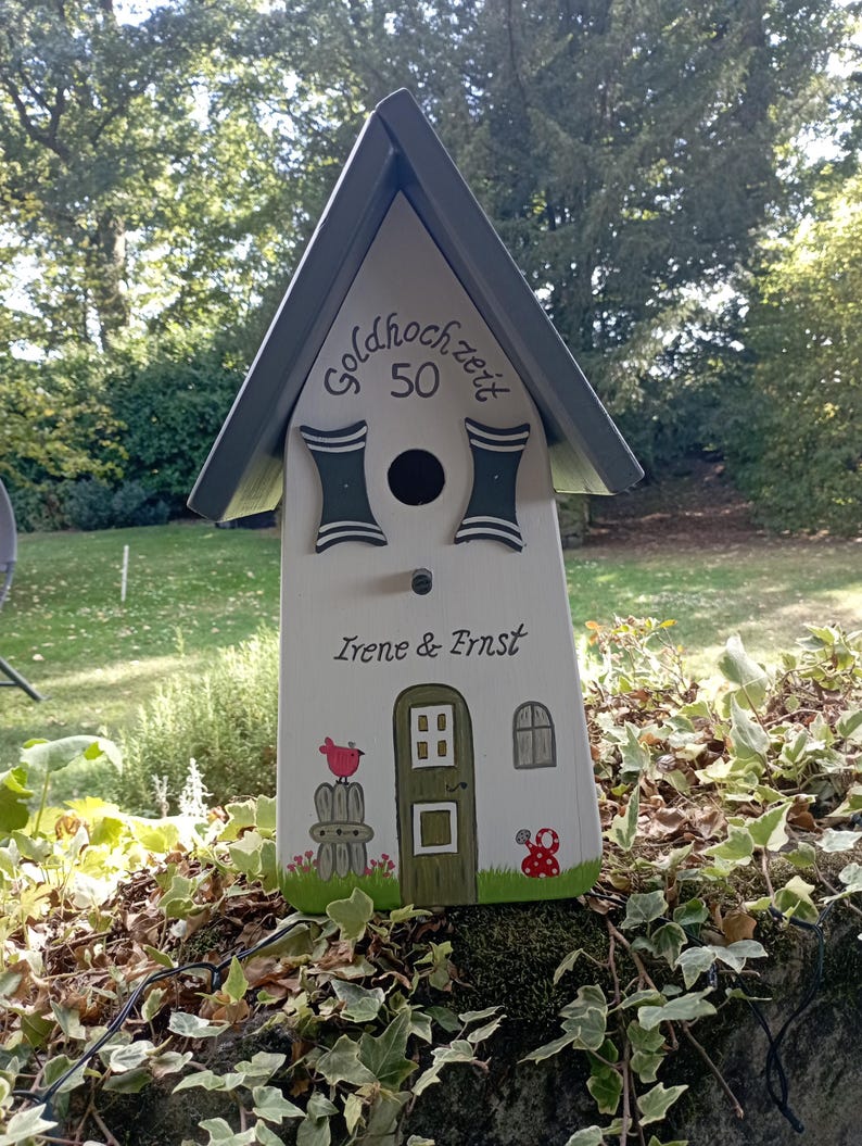 May include: A decorative birdhouse with a gray roof and white body. The birdhouse has the text "Goldhochzeit 50" and the names "Irene & Ernst". It features painted details like a door, window, and a red bird. The birdhouse is set against a backdrop of greenery.