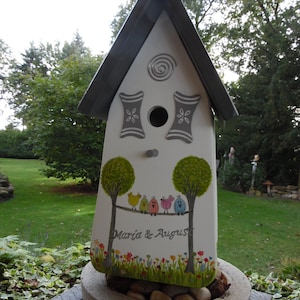 Puede incluir: Una pajarera blanca con techo gris y detalles pintados. La pajarera presenta un dise&ntilde;o de &aacute;rboles, p&aacute;jaros coloridos y los nombres "Maria & August". Se encuentra en una jardinera de piedra llena de guijarros.