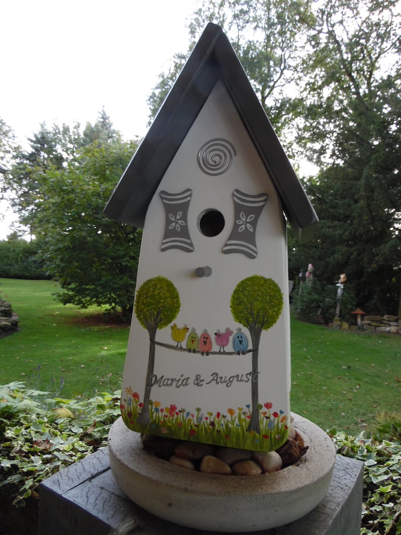 Puede incluir: Una pajarera blanca con techo gris y detalles pintados. La pajarera presenta un dise&ntilde;o de &aacute;rboles, p&aacute;jaros coloridos y los nombres "Maria & August". Se encuentra en una jardinera de piedra llena de guijarros.