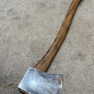 May include: Axe with a wooden handle and a silver-colored blade. The handle is curved and made of a light brown wood. The axe head is a shiny silver color.