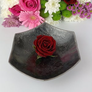 May include: A dark grey metal bowl with a textured surface, featuring a single red rose in the centre. The bowl is surrounded by a bouquet of pink, white and red roses.