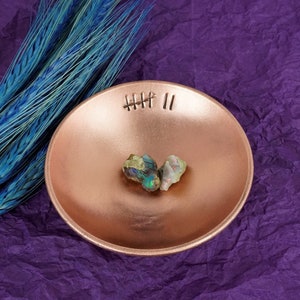 May include: Two iridescent opal stones in a copper bowl with a decorative etched design. The bowl is sitting on a purple textured surface.