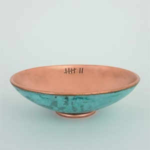 May include: A shallow bowl with a metallic copper interior and a textured turquoise exterior. The inner rim features a stamped tally mark design consisting of five horizontal lines crossed by a diagonal line, followed by two vertical lines.