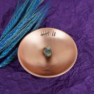 May include: A copper-colored bowl with a small iridescent stone in the center. The bowl has a stitched design with the text "HHH II" on the rim. The bowl is sitting on a purple textured surface.