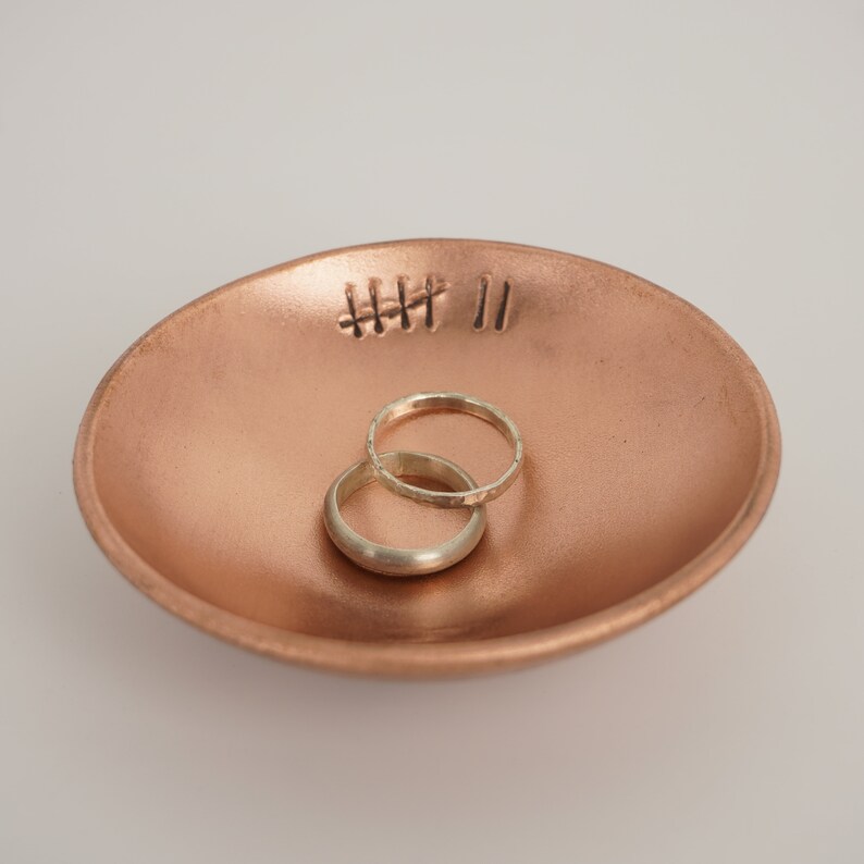 May include: A copper ring dish with two silver rings inside. The dish has a design of four lines and two parallel lines etched into the surface.