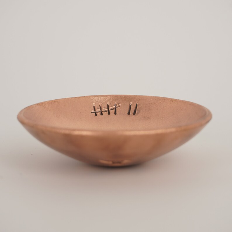 May include: A small, copper-colored dish with a slightly rounded bottom. The dish has a simple design with a small, engraved pattern of lines on the inside.