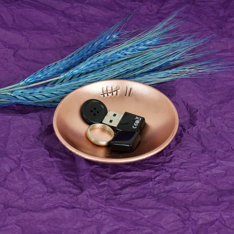 May include: A small, rose gold colored dish with a stitched design and the numbers "III II" on the inside. The dish contains a black button, a silver ring, and a black USB drive.