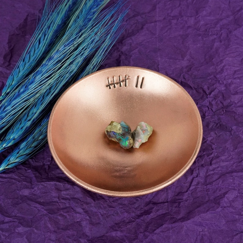May include: Two iridescent opal stones in a copper bowl with a decorative etched design. The bowl is sitting on a purple textured surface.