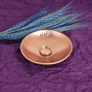 May include: A small, rose gold colored ceramic ring dish with a ring inside. The dish has the words "HHH II" etched into the surface. The dish is sitting on a purple textured surface.