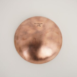May include: A copper-colored bowl with a hammered finish. The bowl has a round shape and a slightly raised rim. The bottom of the bowl has a small, stamped mark that reads "HAND MADE".