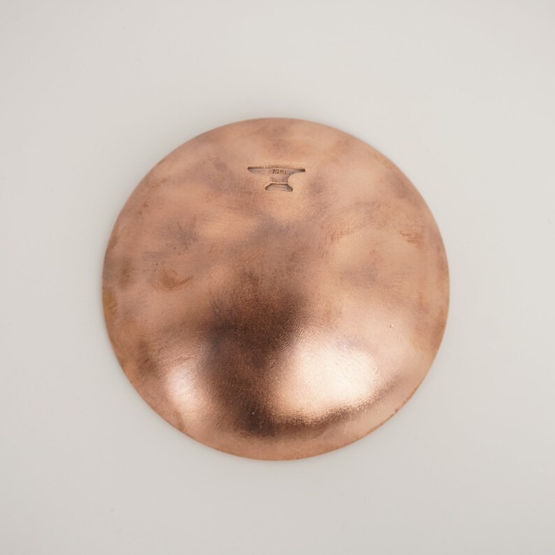 May include: A copper-colored bowl with a hammered finish. The bowl has a round shape and a slightly raised rim. The bottom of the bowl has a small, stamped mark that reads "HAND MADE".
