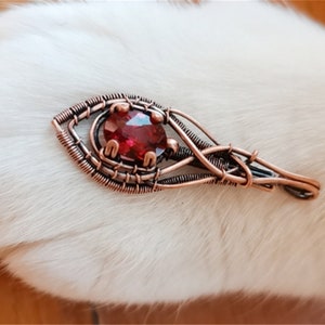 Könnte beinhalten: Ein Anhänger aus umwickeltem Kupferdraht mit einem roten Edelstein in der Mitte. Der Anhänger hat die Form eines Auges und ein filigranes, kompliziertes Design.