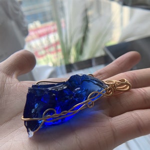 May include: A deep blue glass pendant wrapped in gold wire. The pendant has a rough, irregular shape and is suspended from a gold wire loop.