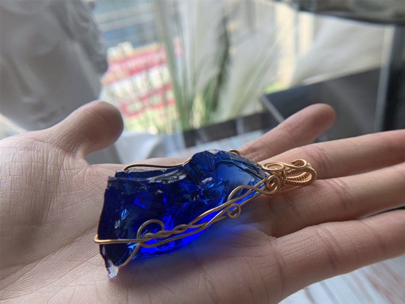 May include: A deep blue glass pendant wrapped in gold wire. The pendant has a rough, irregular shape and is suspended from a gold wire loop.