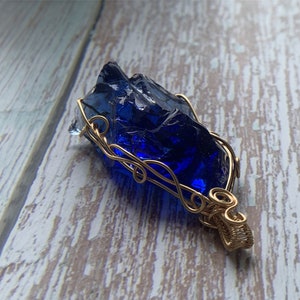 May include: A deep blue glass pendant wrapped in gold wire. The pendant has a rough, irregular shape, resembling a natural crystal.