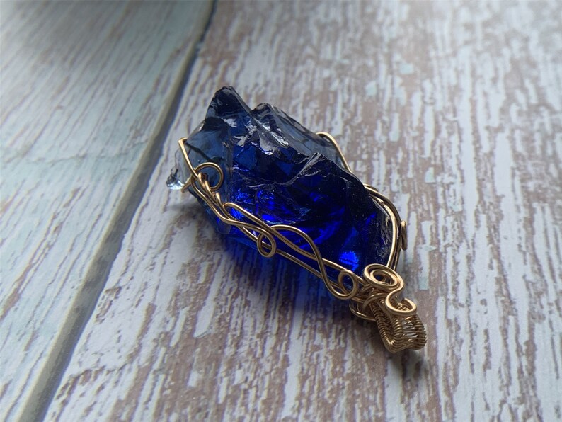 May include: A deep blue glass pendant wrapped in gold wire. The pendant has a rough, irregular shape, resembling a natural crystal.