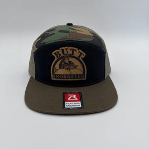 May include: A black, green and brown camouflage trucker hat with a black patch that says "BUTT SNORKELER" in white letters. The patch features a cartoon image of a person wearing a snorkel and swimming in a pool of water. The hat is made by Richardson and has a flat bill.