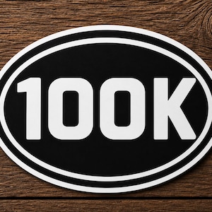 May include: An oval-shaped sticker or sign featuring a black background with a white border and the bold white text 100K centered in the middle. The design is clean and high-contrast, displaying the number clearly in a sans-serif font style.