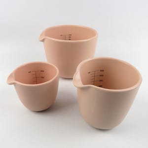 Op de afbeelding: Drie perzik kleurige maatbekers van verschillende formaten. Elke beker heeft een schenktuit en zwarte meetmarkeringen in milliliters. De bekers zijn op een witte ondergrond geplaatst.
