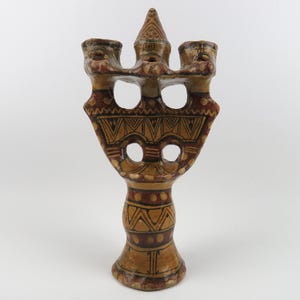 May include: A hand-painted ceramic candelabra featuring a tan base color with dark brown and red geometric patterns. The structure has a central spire flanked by two candle holders, with open circular cutouts and intricate triangular line work.