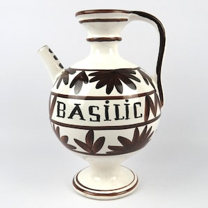 Puede incluir: Jarra de cerámica blanca con asa, boquilla y detalles decorativos marrones. La palabra "BASILIC" está impresa en marrón en el cuerpo, adornada con motivos florales marrones. La jarra tiene una base de pedestal.