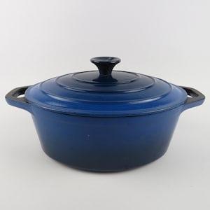 Peut inclure: Une cocotte en fonte émaillée bleu profond avec son couvercle assorti. La marmite présente deux poignées latérales intégrées et un bouton noir central sur le couvercle. L'extérieur a une finition lisse et brillante avec un dégradé de bleu.