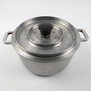 Könnte beinhalten: Ein silberfarbener Kochtopf aus Metall mit Deckel und zwei Griffen. Der Deckel hat einen schwarzen Knopf und das Wort "TOURUS" ist sichtbar. Der Topf ist rund und scheint aus Aluminium zu sein.