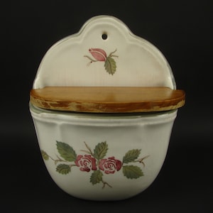 May include: A white ceramic wall-mounted salt box with a wooden shelf and lid. The box is decorated with red roses and green leaves. A single rose is painted on the top. The box has a small hole for hanging.