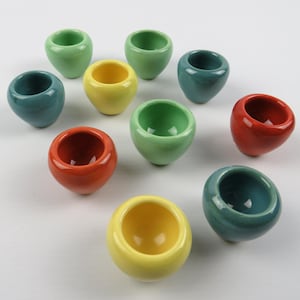 Peut inclure: Une collection de onze petits bols en céramique de différentes couleurs, dont vert, jaune, rouge et sarcelle. Les bols ont une forme arrondie et une finition lisse et brillante. Ils sont disposés sur une surface blanche.