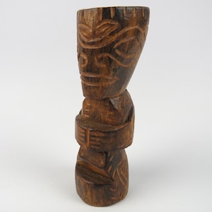 Könnte beinhalten: Eine geschnitzte Tiki-Statue aus Holz mit dunkler brauner Oberfläche. Die Statue zeigt ein stilisiertes Gesicht mit geschnitzten Details und einem gestapelten Design. Der Tiki ist ein dekorativer Gegenstand, wahrscheinlich für die Inneneinrichtung oder eine thematische Umgebung.
