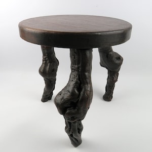 May include: A dark brown, circular side table with three distinctive legs. The tabletop is a solid, dark brown. The legs have a textured, organic appearance, giving the table a rustic, natural aesthetic. The table's diameter is approximately 40 cm.
