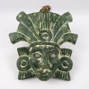 Op de afbeelding: Een groene, verweerde masker met een gedetailleerd gezicht en hoofdtooi. Het masker heeft oog- en mondopeningen en cirkelvormige oorversieringen. Een bruin touwtje is aan de bovenkant bevestigd om op te hangen. Het masker is waarschijnlijk een decoratief item.