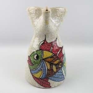 Może przedstawiać: Ceramiczny dzban z motywem ryby. Dzban jest beżowy z teksturowaną powierzchnią i dzióbkiem. Kolorowa ilustracja ryby z zielonymi, żółtymi, czerwonymi i niebieskimi łuskami jest namalowana z przodu. Dzban prawdopodobnie służy do serwowania napojów.