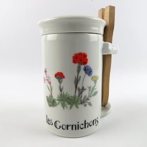 May include: White ceramic jar with lid, featuring a floral design with red, blue, and yellow flowers. The jar has a handle and a wooden spoon is attached. The text "les Cornichons" is printed on the jar.