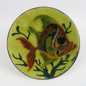 May include: A ceramic plate with a hand-painted fish design. The fish is brown, orange, and black, set against a light green background with dark blue seaweed accents. The plate has a black rim and a glossy finish.