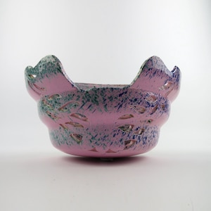 Peut inclure: Un bol en céramique avec une base rose ornée d'un motif tacheté de éclaboussures de peinture bleue et verte. Le bol présente un bord festonné et des ouvertures décoratives en forme de feuilles sur les côtés, créant un design ajouré.