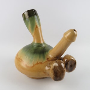 May include: A ceramic sculpture in the shape of a phallus. The sculpture features a bulbous base with a green and tan glaze, a long shaft, and two rounded forms. The sculpture is tan and brown.
