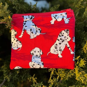May include: A red zippered pouch featuring a repeating pattern of white and black dalmatian dogs. The dogs are in various poses against a solid red background. The zipper is red.