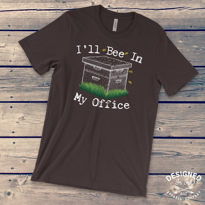 May include: A dark brown t-shirt with a white graphic of a beehive and the text "I'll Bee In My Office".