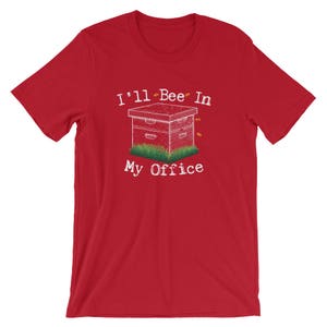May include: Red short-sleeved t-shirt with white text that reads "I'll Bee In My Office". The text is accompanied by a black and white illustration of a beehive with bees flying around it.