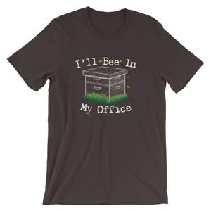 May include: A dark gray t-shirt with the text "I'll Bee In My Office" in white. A cartoon beehive with bees flying around it is below the text.
