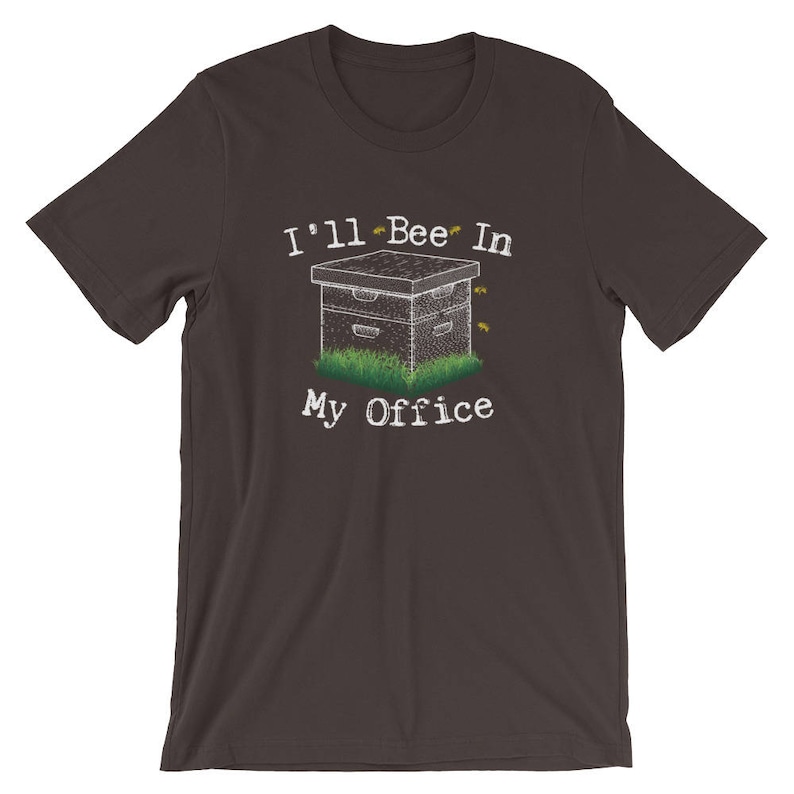 May include: A dark gray t-shirt with the text "I'll Bee In My Office" in white. A cartoon beehive with bees flying around it is below the text.