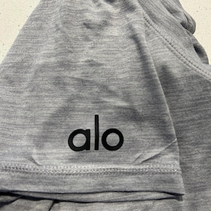 May include: A heather grey short-sleeved t-shirt featuring the black lowercase text "alo" printed on the sleeve. The garment is constructed from a soft, heathered fabric with visible stitching along the hem and sleeve seams.
