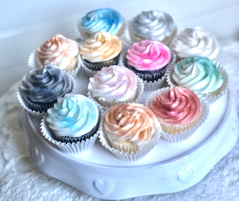 May include: A white cake stand with 12 cupcakes decorated with different colored frosting. The frosting is swirled and has a watercolor effect. The colors include blue, pink, gray, gold, and silver.