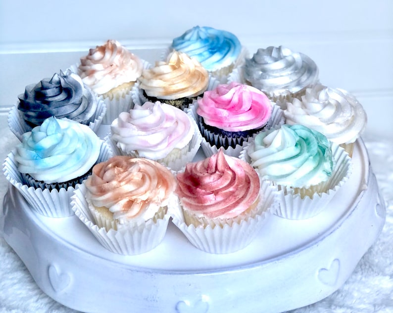May include: A white cake stand with a variety of cupcakes decorated with different colored frosting. The cupcakes are decorated with shades of blue, pink, gray, and brown.