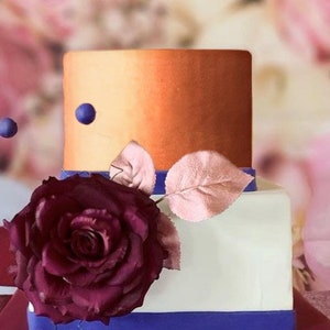 May include: A two-tiered wedding cake with a white bottom tier and a top tier with an ombre orange and gold finish. The cake is decorated with a single large burgundy rose and a few small blue beads.