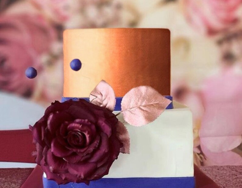 May include: A two-tiered wedding cake with a white bottom tier and a top tier with an ombre orange and gold finish. The cake is decorated with a single large burgundy rose and a few small blue beads.